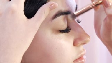 Kız stilisti stüdyodaki genç ve güzel bir kadına şık, parlak bir makyaj yapıyor. Özel bir fırçayla göz farı uygular