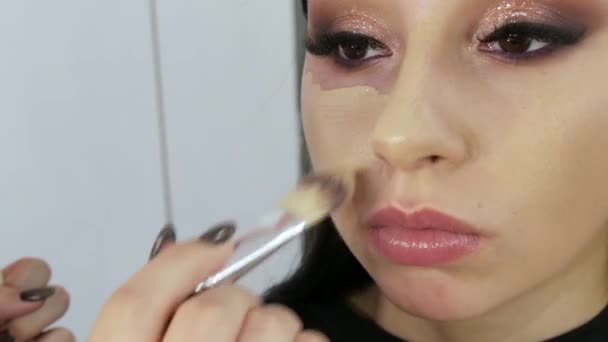 Pincel aplica fundação para o rosto de um belo modelo jovem para maquiagem à noite olhos fumegantes — Vídeo de Stock