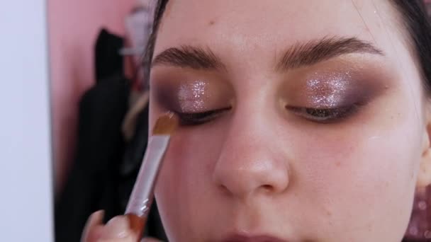 Estilista maquillaje maestro hace un ojo en la noche elegante maquillaje brillante en un modelo de chica joven en un estudio de belleza — Vídeos de Stock