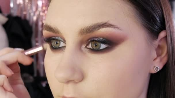 Beautiful expressive eyes of a young female model with bright smoky eyes makeup. A special makeup brush applies foundation or concealer under the eyes — Stock Video
