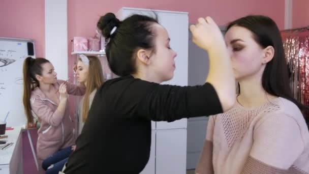 23 de março de 2021 - Kamenskoe, Ucrânia: artista de maquiagem para meninas aplica maquiagem no rosto do modelo na frente do espelho no estúdio de beleza. Mesa com paletas de cosméticos, batons, escovas, fundação — Vídeo de Stock