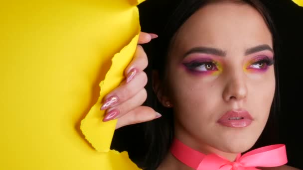 Una bella modella giovane ragazza con un brillante trucco di moda giallo succoso e un fiocco rosa intorno al collo su uno sfondo di elegante carta gialla. Immagine modello di moda — Video Stock