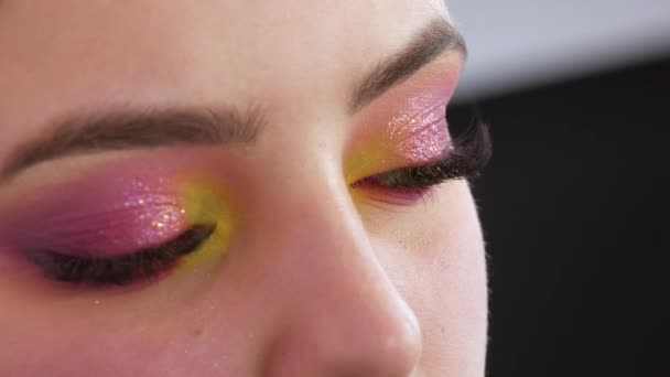 Artista de maquillaje profesional hace maquillaje de moda brillante para modelar en sombras de ojos amarillos y rosados en los párpados en el estudio, vista de cerca — Vídeos de Stock