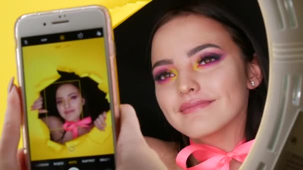 Um belo modelo de menina com uma maquiagem de moda amarela suculenta brilhante e um arco rosa em torno de seu pescoço em um fundo de papel amarelo elegante. Imagem de modelo de moda. Idioma russo no menu do telefone — Vídeo de Stock