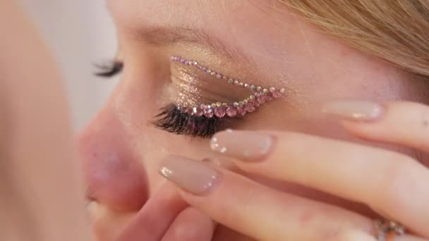 Seorang seniman tata rias profesional menerapkan rhinestone khusus untuk mata dan kelopak mata seorang model gadis cantik muda. Berdandan ala fashion — Stok Video