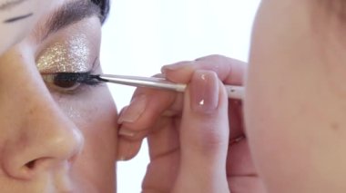 Kız stilisti makyaj sanatçısı, siyah göz kaleminin ince bir çizgisini model gözündeki altın tonlarda parlak makyaja, yakın çekim görüntüsüne uyguluyor.