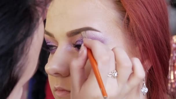 Een professionele make-up kunstenaar met een speciale borstel corrigeert en schildert de vorm van de wenkbrauwen van een jong mooi meisje model. Schitterende mode make-up — Stockvideo