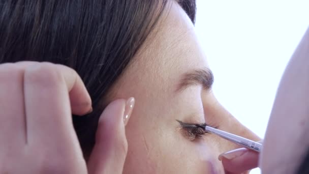 Девушка стилист макияж художник наносит тонкую линию черной подводки для глаз на блестящие макияж в золотых тонах на модели глаз, крупным планом — стоковое видео