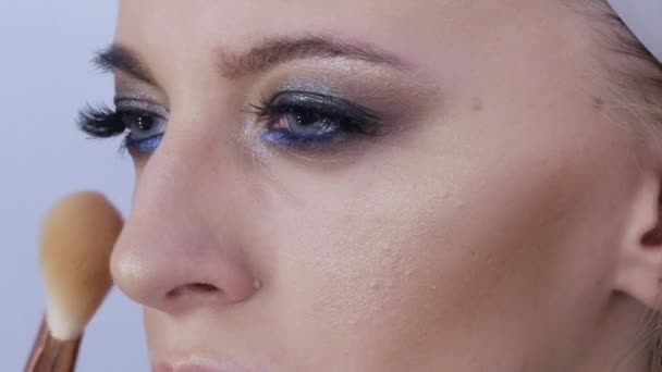 Cepillo especial para empolvar la cara. Un artista de maquillaje profesional hace un maquillaje de noche brillante para los ojos ahumados en un modelo con ojos azules, vista de cerca — Vídeos de Stock