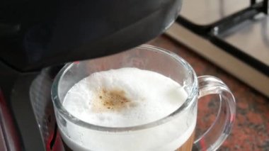 Özel bir kapsül kahve makinesinde sabah kahvesi sıcak bir içecek yapar. Ev mutfağındaki şeffaf cam bardakta süt ve kahve tabakları.
