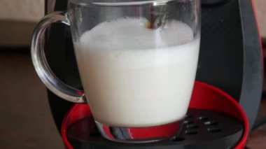 Özel bir kapsül kahve makinesinde sabah kahvesi sıcak bir içecek yapar. Ev mutfağındaki şeffaf cam bardağa sıcak süt dökülüyor.
