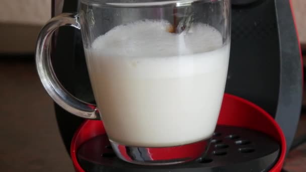 Morgendlicher Latte in einer speziellen Kapselkaffeemaschine macht ein Heißgetränk. Heiße Milch wird in der heimischen Küche in einen transparenten Glasbecher gegossen — Stockvideo