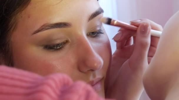 Um artista de maquiagem profissional aplica uma sombra brilhante em uma sombra nua com um pincel especial para uma jovem modelo linda — Vídeo de Stock
