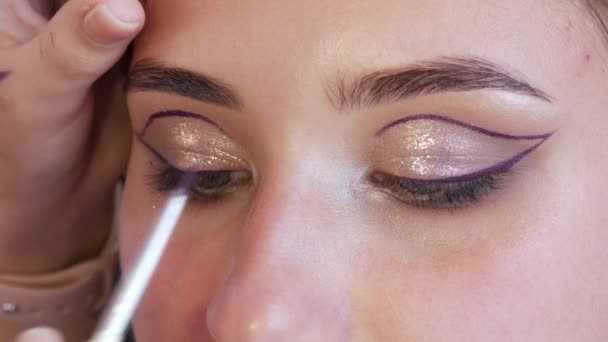 Girl stylist make-up artist menerapkan garis tipis eyeliner ungu pada make-up mengkilap dalam nada emas pada model mata, close-up view — Stok Video
