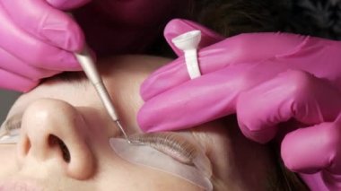 Yetişkin bir kadın, profesyonel bir güzellik salonunda modern kirpik temizleme prosedürüyle karşı karşıya. Pembe lastik eldivenlerle kirpik kıvırma prosedüründen önce usta özel yapıştırıcı uyguluyor.