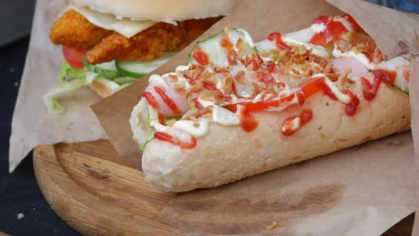 Pyszny duży hot dog z dużą ilością sosu majonezowego, smażona cebula, kiełbasa, ketchup, ogórek na ladzie festiwalu ulicznego jedzenia — Wideo stockowe