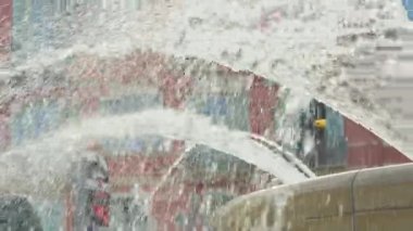 Şehir çeşmesinden yansıyan dinamik su akıntıları ve güneş ışığı yansımaları. Soyut hareket ve berrak su dokusu ve arka planda renkli binalar. Kentsel yaz atmosferi tazelik ve hareketle dolu