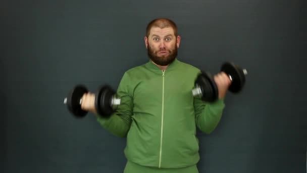 Um homem com barba num fato de treino levanta as mãos com halteres de ferro pesados. — Vídeo de Stock