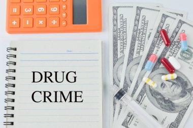 İlaç kapsülleri, şırınga, para banknotları, hesap makinesi ve DRUG CRIME ile yazılmış defter.