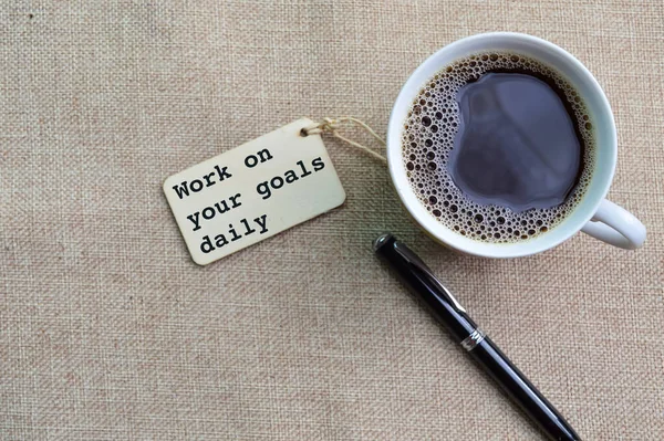 A cup of coffee, pen and label tag written with text WORK ON YOUR GOALS DAILY