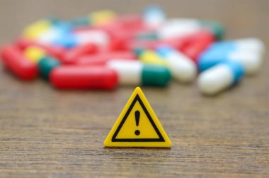 Üzerinde ünlem işareti olan sarı bir uyarı işareti, hapların bulanık arka planına karşı, ilaç risklerini ve uyuşturucu güvenliğini vurguluyor..