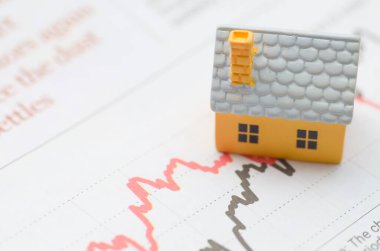 Konut Piyasası Değişkenliği: Değişken bir finans grafiğindeki minyatür bir ev, değişen emlak piyasası ve yatırım risklerini gösteriyor.