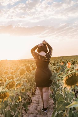A beautiful young girl with her back turned touching her hair and walking through a field of sunflowers in a sunset of a summer day