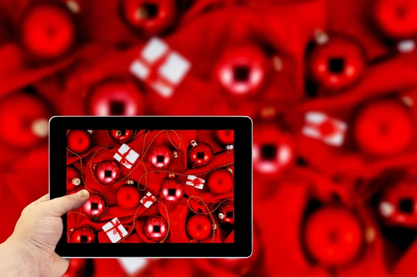 Tablet-Fotografie. Fotografieren auf einem Tablet. Hintergrund aus roten Christbaumkugeln, kleinen Geschenkschachteln und Goldschmuck auf rot glänzendem Seidenstoff — Stockfoto