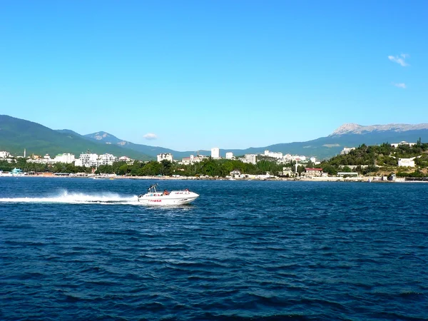 Alushta, Crimea, Ucrania - 27 de junio de 2009: Lancha blanca flotante en el fondo de la ciudad en primera línea de mar con gran montaña —  Fotos de Stock