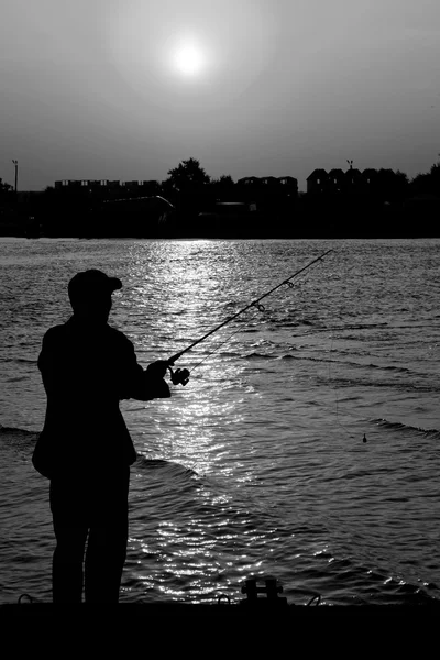 Pescador de pé na borda da doca com vara de pesca perto do rio e cidade de praia em raios de pôr do sol preto e branco — Fotografia de Stock