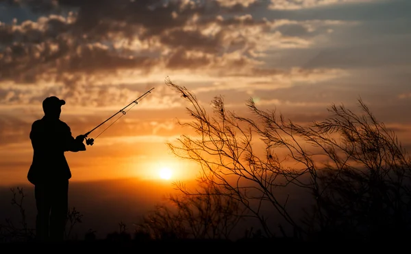 Fischer, der mit Angelrute in der Nähe des Flusses auf dem Hintergrund von Pflanzen und schönen bewölkten Himmel mit goldenem Sonnenuntergang und Lichtstrahlen steht — Stockfoto