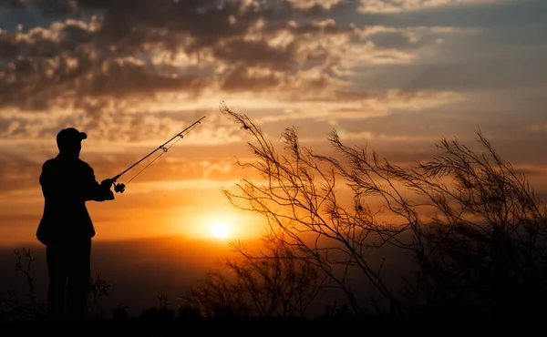 Fisherman standing with fishing rod near river on background of plants and beautiful cloudy sky with golden sunset and rays of light — Stock Photo, Image