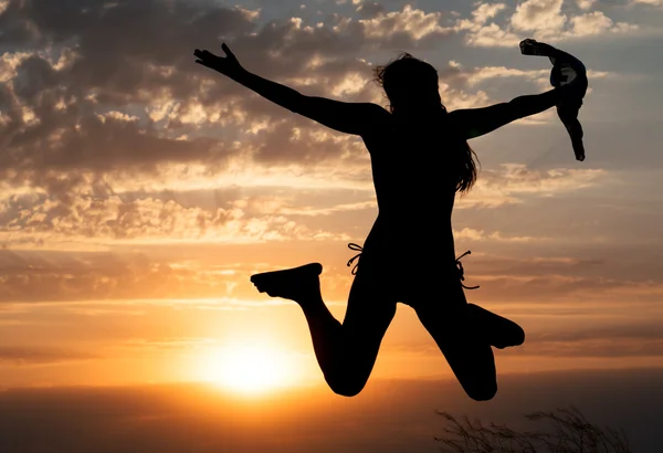 Chica joven saltando silueta con chal sobre fondo de hermoso cielo nublado con puesta de sol naranja y rayos de luz —  Fotos de Stock