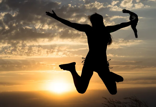 Chica joven saltando silueta con chal sobre fondo de hermoso cielo nublado con puesta de sol amarillo y rayos de luz —  Fotos de Stock