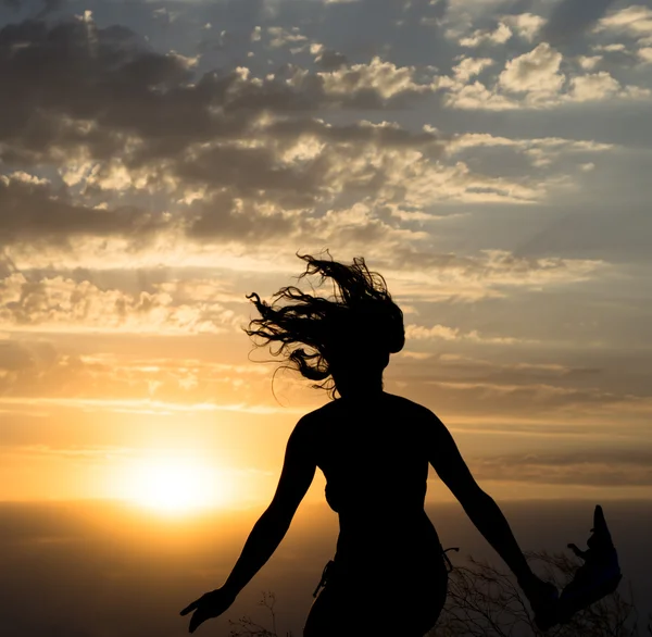 Fiatal lány jumping silhouette kendőt, a háttérben a gyönyörű felhős ég a sárga naplementére és fénysugarak — Stock Fotó