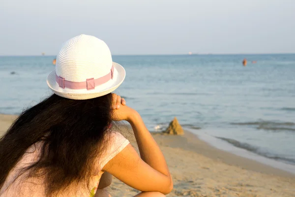 Fille brune en chapeau blanc regardant la mer tout en étant assis sur la plage — Photo