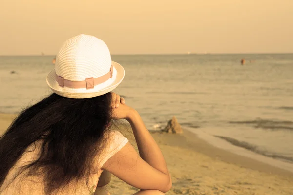 Fille brune en chapeau blanc regardant la mer tout en étant assis sur la plage chaud filtré — Photo