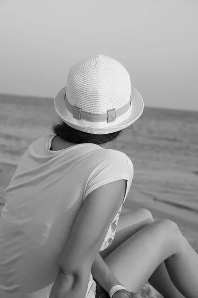 Fille brune en chapeau blanc regardant la mer tout en étant assis sur la plage noir et blanc — Photo