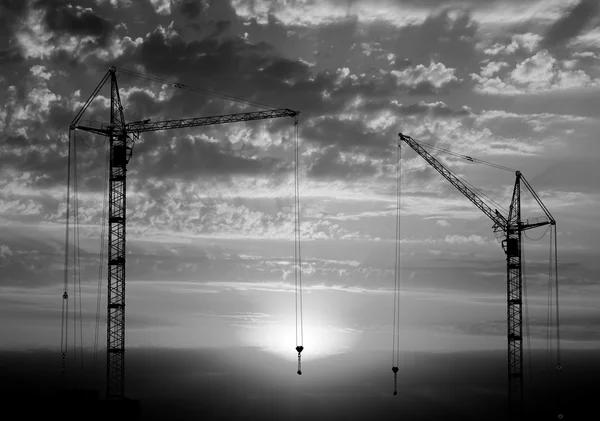 Hoisting cranes working on beautiful cloudy sky with sunset and rays of light background black and white — Stock Photo, Image