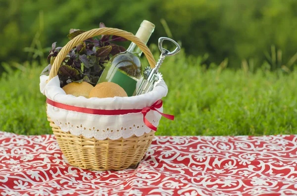 Cesta de picnic decorada con una botella de vino blanco, sacacorchos, bollos y manojo de albahaca en mantel rojo, paisaje verde Imagen de stock