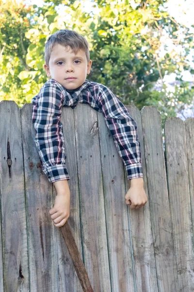 El chico de campo con camisa a cuadros se sienta en una valla. Concepto de actividad infantil. — Foto de Stock