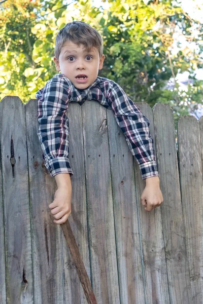 O rapaz do campo de camisa quadriculada senta-se numa cerca. Conceito atividade infantil. — Fotografia de Stock