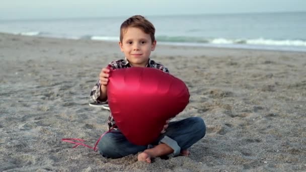 Lindo chico se sienta en la playa y abraza un globo rojo del corazón. — Vídeo de stock