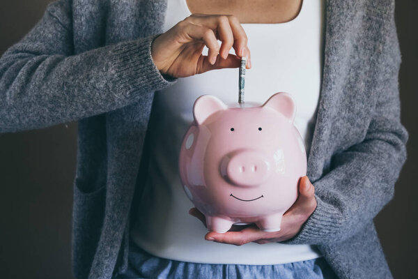 Close-up young woman putting bill in piggy bank. Female saving money for household payments, bank bills, calculating monthly family budgets, making investments or strategy for retirement.