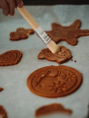 Kek tepsisinde zencefilli kurabiyeleri parlatan bir fırçanın yakın çekimi. Noel pişirmeye hazırlanan, sıcak, ev yapımı tatil atmosferini yakalayan şenlikli şekillerin ayrıntılı görüntüsü.