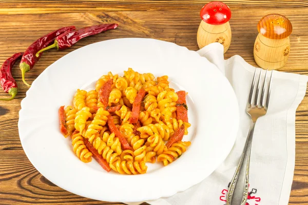Italian pasta fusilli with tomato sauce and sausage in plate, napkin, fork, pepper on wooden table — Zdjęcie stockowe