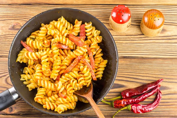 Italian pasta fusilli with tomato sauce and sausage in pan, wooden spoon, red pepper on table, top view, space for text — Zdjęcie stockowe