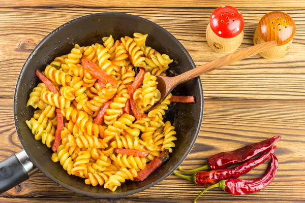 Italian pasta fusilli with tomato sauce and sausage in pan, wooden spoon, red pepper on table, top view, space for text — Zdjęcie stockowe
