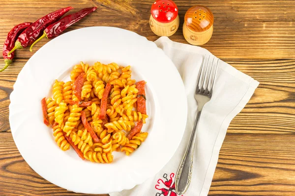 Italian pasta fusilli with tomato sauce and sausage in plate, napkin, fork, pepper on wooden table, top view — Zdjęcie stockowe