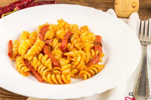 Italian pasta fusilli with tomato sauce and sausage in plate, napkin, fork on wooden table — Zdjęcie stockowe